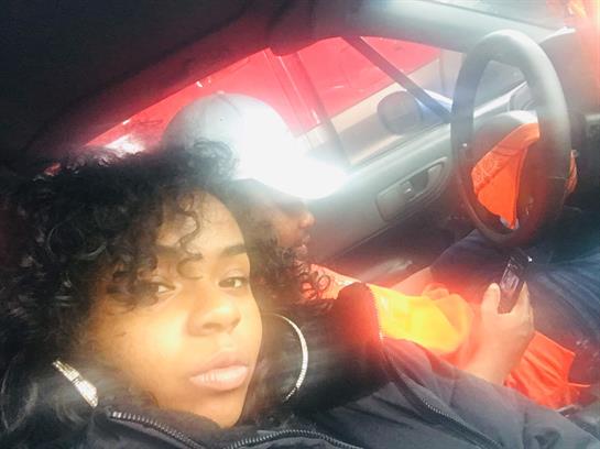 A young woman relaxes in a car, dressed in vibrant clothes against a striking backdrop.