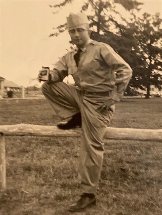 A soldier casually poses on a fence while holding a drink in a quiet location.