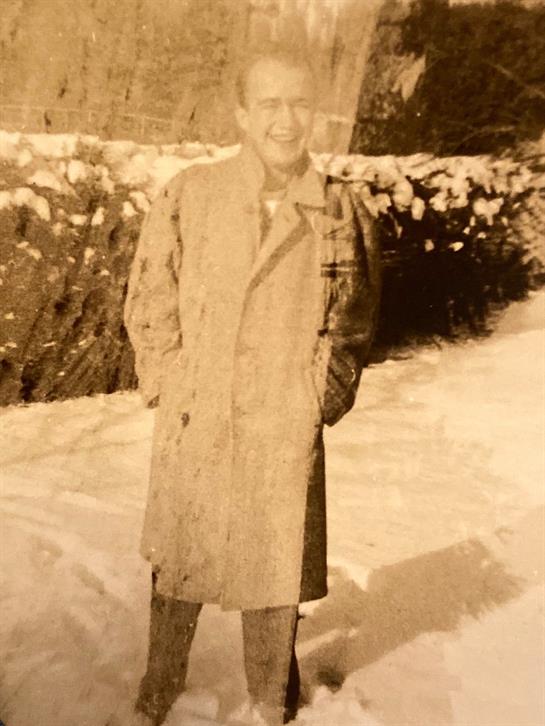 A man in a light coat stands in a snowy landscape, smiling amid the winter chill.