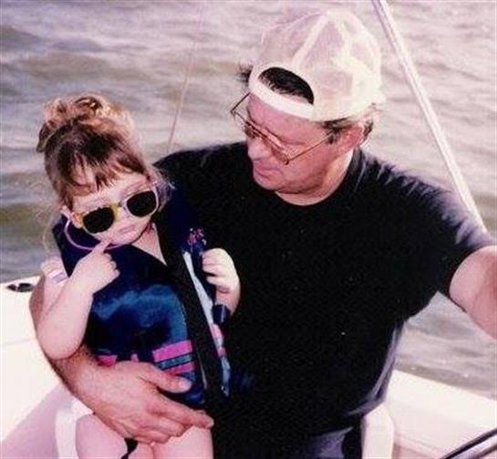 A young child in sunglasses is seated on an adult's lap while enjoying a boat ride.