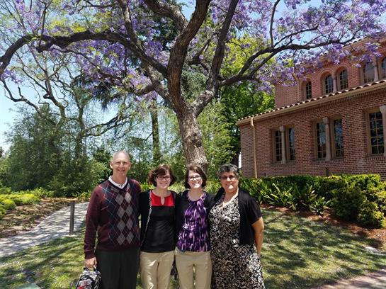 Four individuals gather joyfully beneath a vibrant jacaranda tree, enjoying the sunny weather.