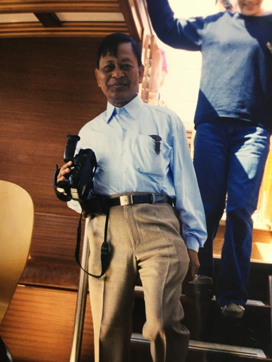 A man stands smiling with a camera in hand, while another person is partially visible behind him.