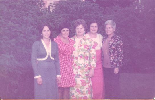 Five women stand closely together wearing vibrant dresses and smiling in a garden setting.