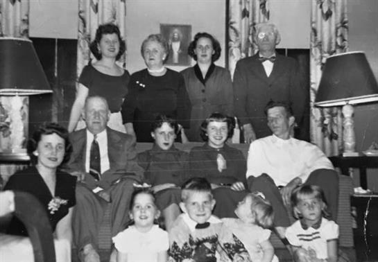 A large group of adults and children poses together in a cozy living room from the 1950s.