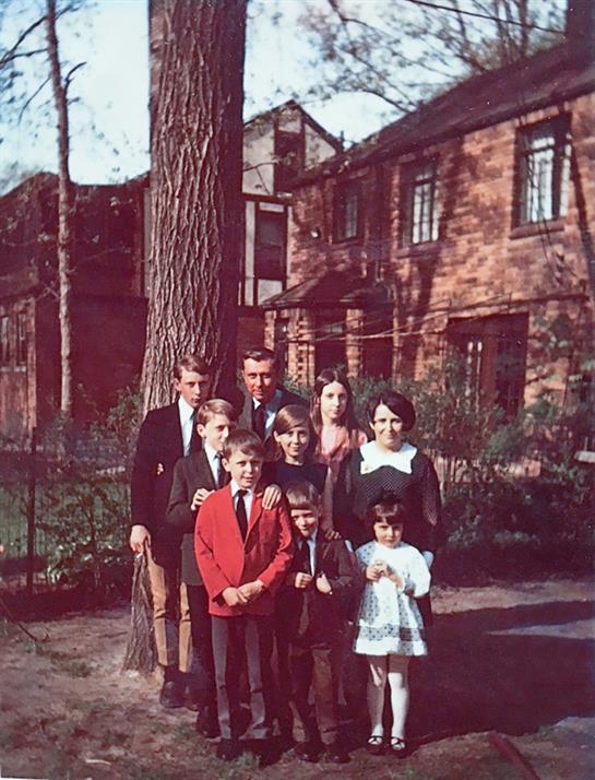 A large family gathers on a front lawn, smiling together in front of their brick house.