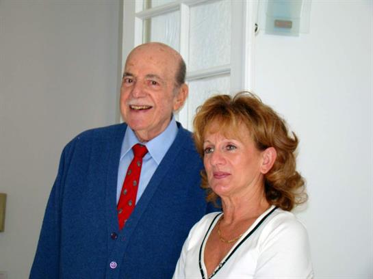 An elderly couple stands side by side, smiling warmly in a well-lit indoor space.