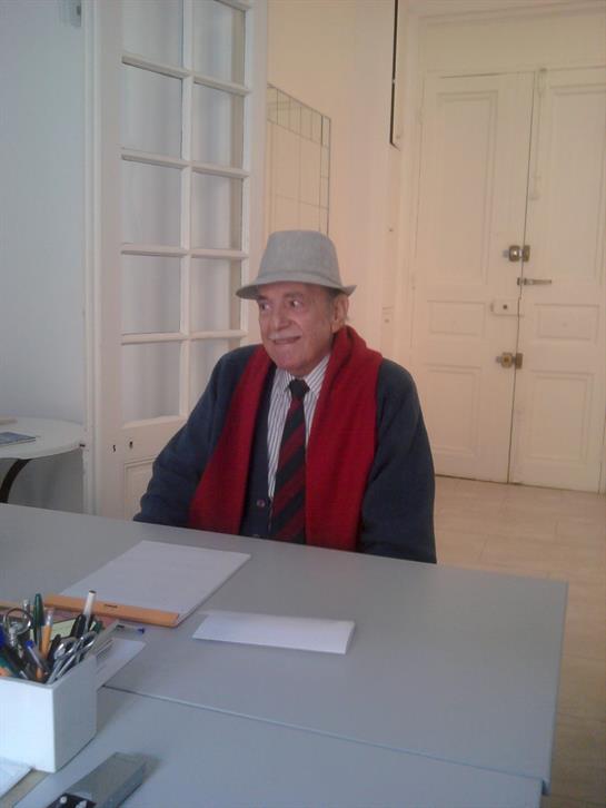 A man in a hat and scarf sits at a desk, surrounded by papers in a bright room.
