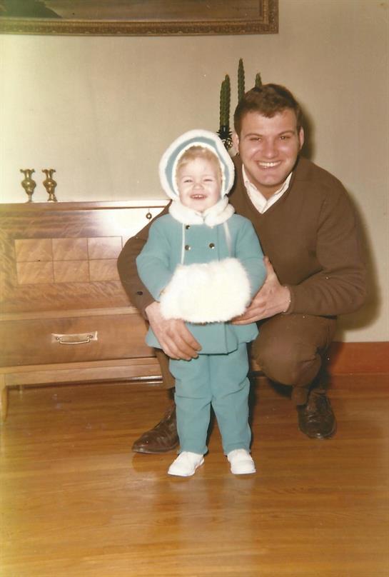 Child beams in warm clothes as father kneels beside, capturing a loving indoor moment.