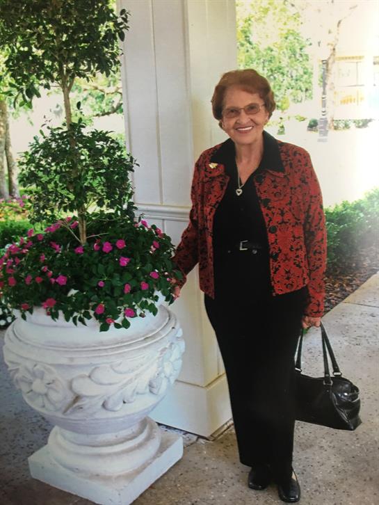 A smiling elderly woman stands next to a decorative planter filled with flowers.