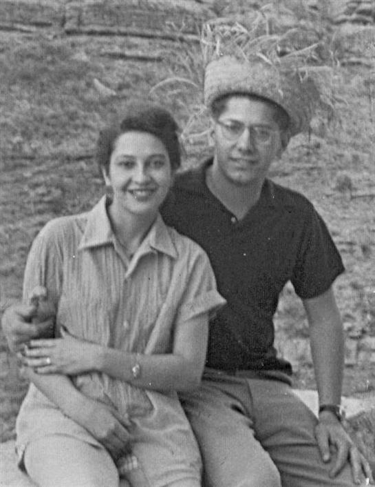 A cheerful couple poses for a casual portrait in a natural landscape, smiling warmly at the camera.