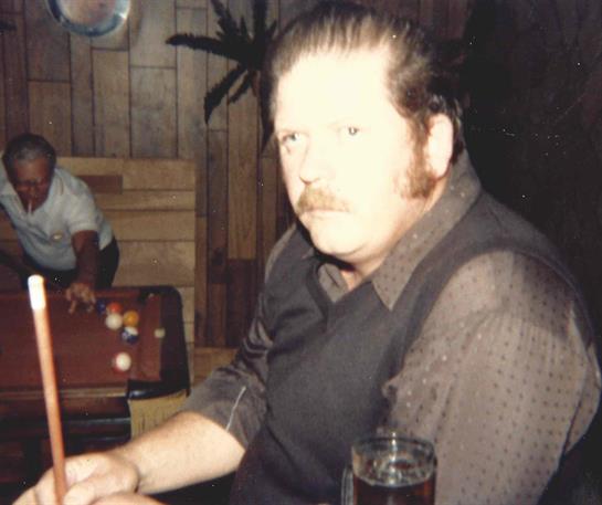 A man with a mustache is seated at the bar, holding a drink and talking with others nearby.