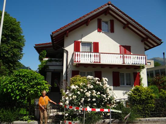 An elderly gentleman poses outside a charming house with vibrant red shutters and a lush garden.