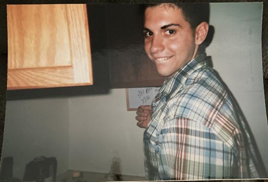Smiling young man in a plaid shirt stands in a kitchen, preparing for a meal during evening.