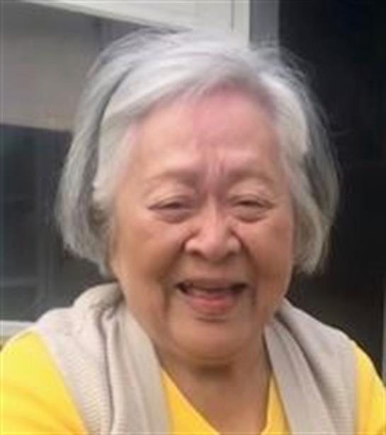 An elderly woman with gray hair smiles joyfully while wearing a yellow shirt on a sunny day.