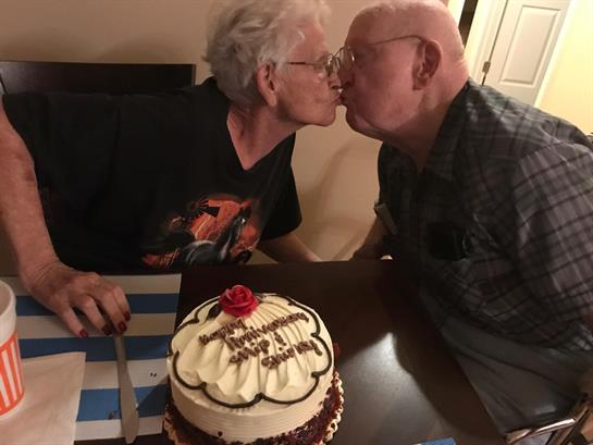Two elderly individuals share a sweet kiss while celebrating a birthday with cake on a table.