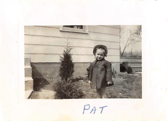Little child stands by a small tree, smiling in a yard with a house in the background.