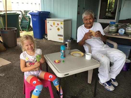 A young girl and an older man share snacks at a table outside, filled with joy and laughter.