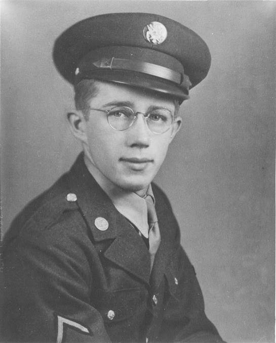 A young soldier wearing glasses and a military uniform sits for a portrait in a studio.