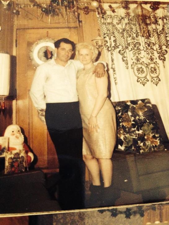 A couple stands closely together in a cozy living room, smiling and dressed in vintage attire.