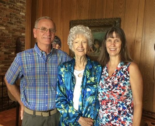 Elderly woman stands between two relatives in a warm wooden room, smiling for the moment.