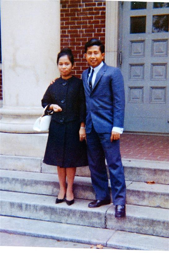 Two individuals dressed in formal clothing stand together on stone steps, smiling.