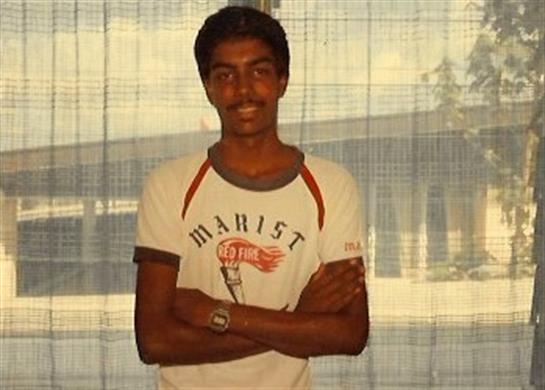 A young man with a friendly smile stands confidently by a window in casual fashion.