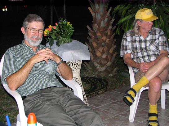 Two men sit comfortably on chairs amidst lush greenery, enjoying a peaceful evening together.