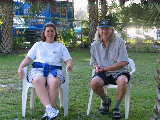 Two individuals sit comfortably in plastic chairs, enjoying a relaxing moment in a lush garden.