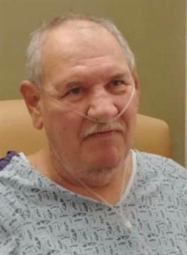 An elderly man in a hospital gown sits thoughtfully in a medical facility with oxygen support.