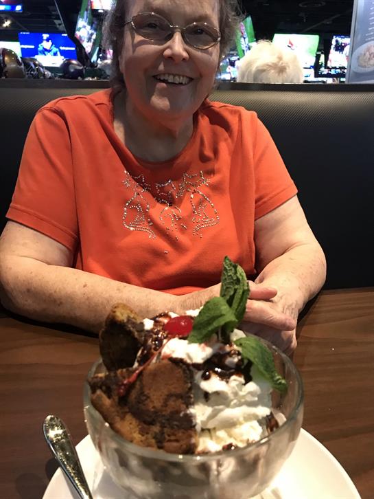 A smiling woman sits at a table, savoring a large sundae topped with cherry and mint.