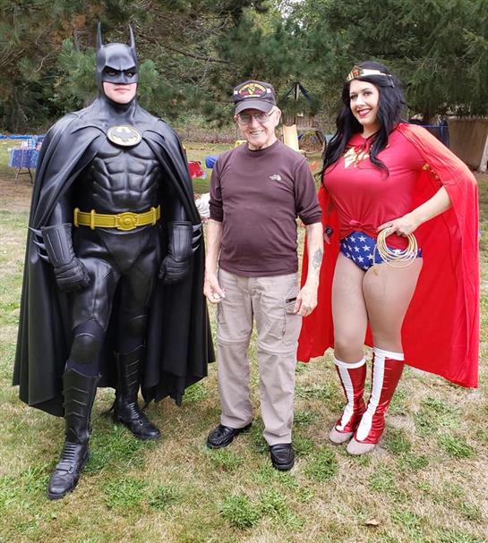 Two costumed superheroes pose with an older man at an outdoor gathering in a park.