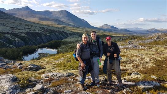 Group of friends poses together on a mountain trail with scenic views and a serene lake nearby.