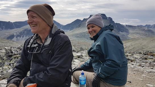 Two friends are sitting on rocks, surrounded by mountains, sharing a joyful moment while hiking.