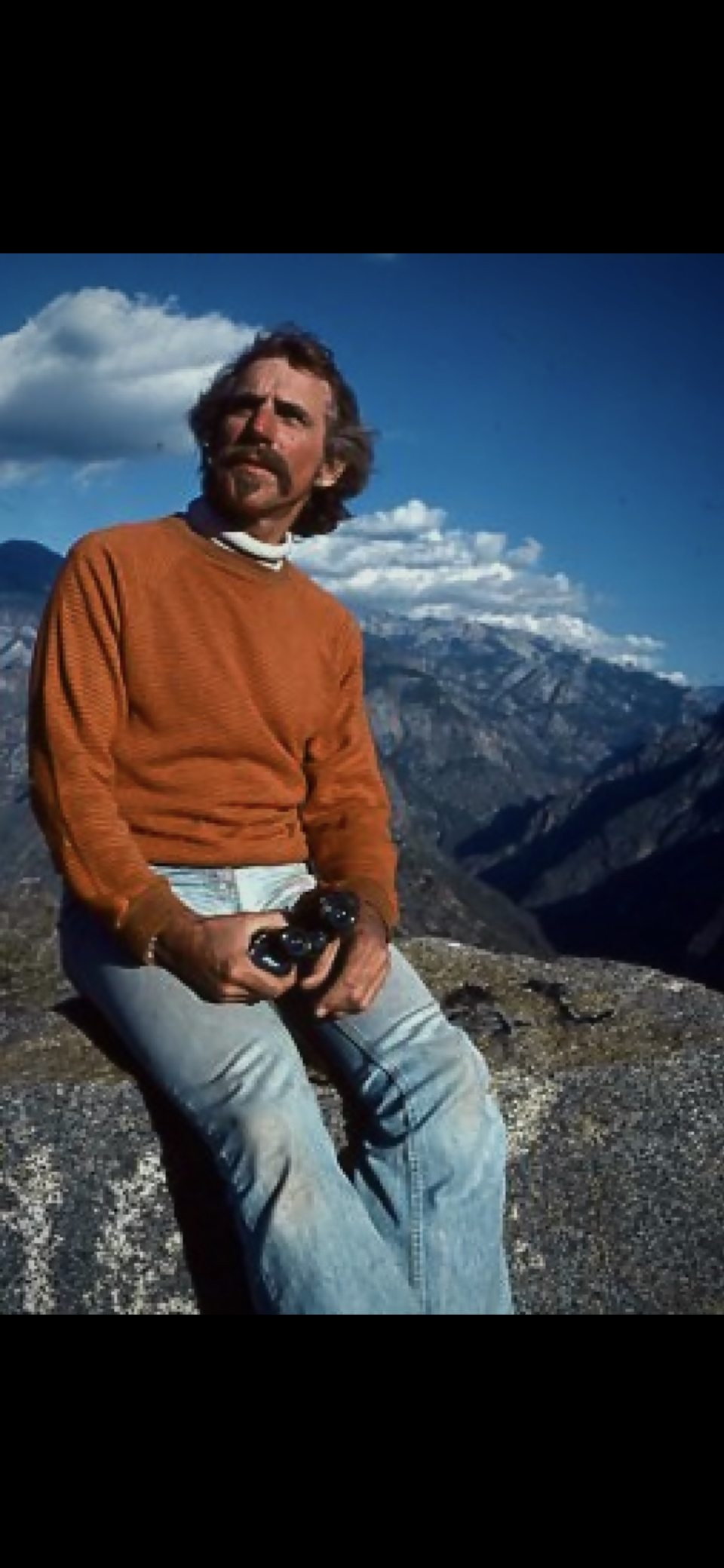 A man in an orange sweater and jeans sits on a rock in a stunning mountain landscape.