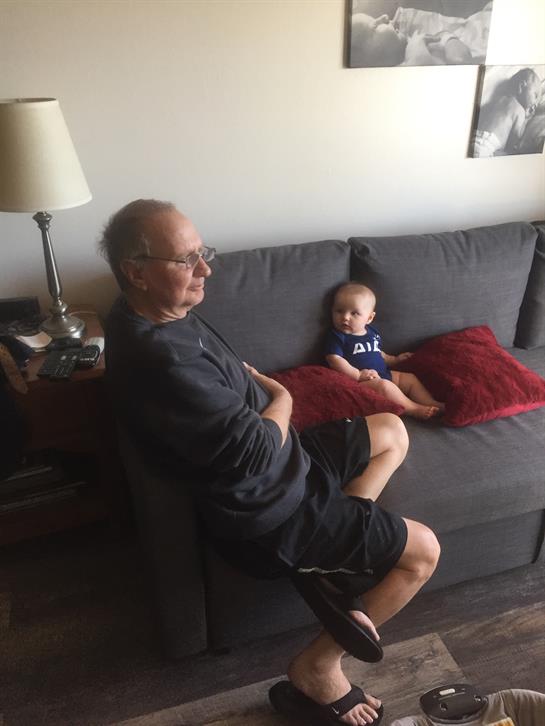 An elderly man sits on a couch while a toddler plays beside him, creating a warm family atmosphere.