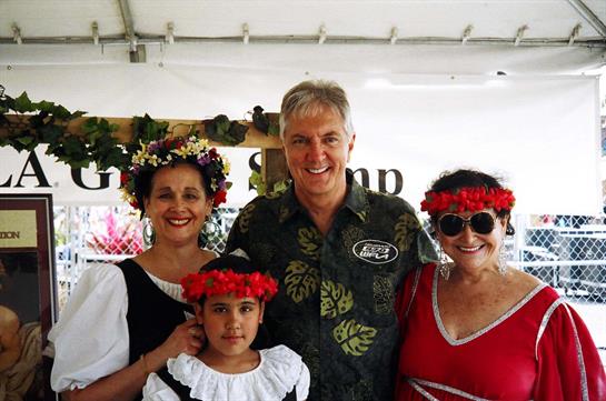 Families gather together at a community festival, dressed in traditional outfits and floral crowns.
