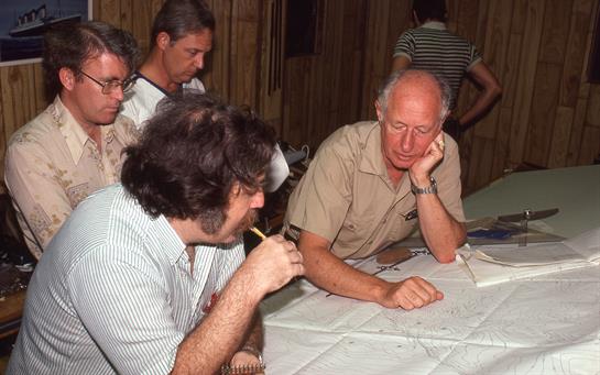 People are gathered around a table studying maps and engaging in serious discussions in the evening.