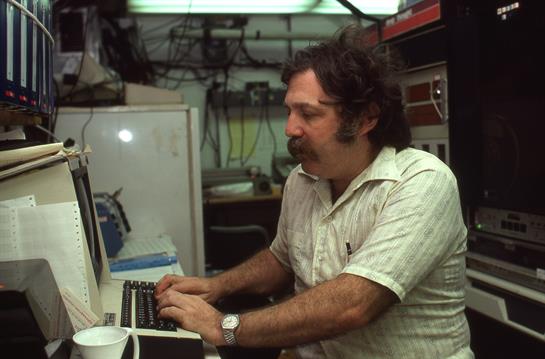 A man sits focused at his computer, typing away while surrounded by equipment and tools.
