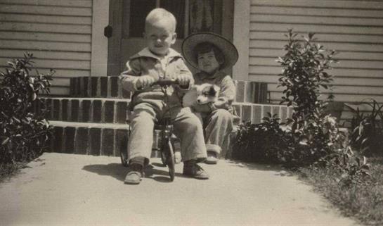 Two boys enjoy a playful moment on a porch, one riding a tricycle while the other cuddles a puppy.