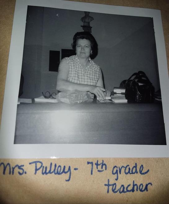 A teacher organizes materials at her desk in a classroom adorned with books, showcasing dedication.