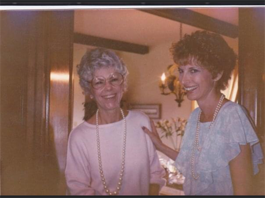 Two women wearing necklaces smile joyfully at a cozy indoor gathering filled with warmth.