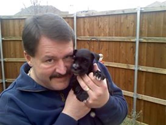 A man enjoys time with a small black puppy, smiling and posing in a fenced backyard.