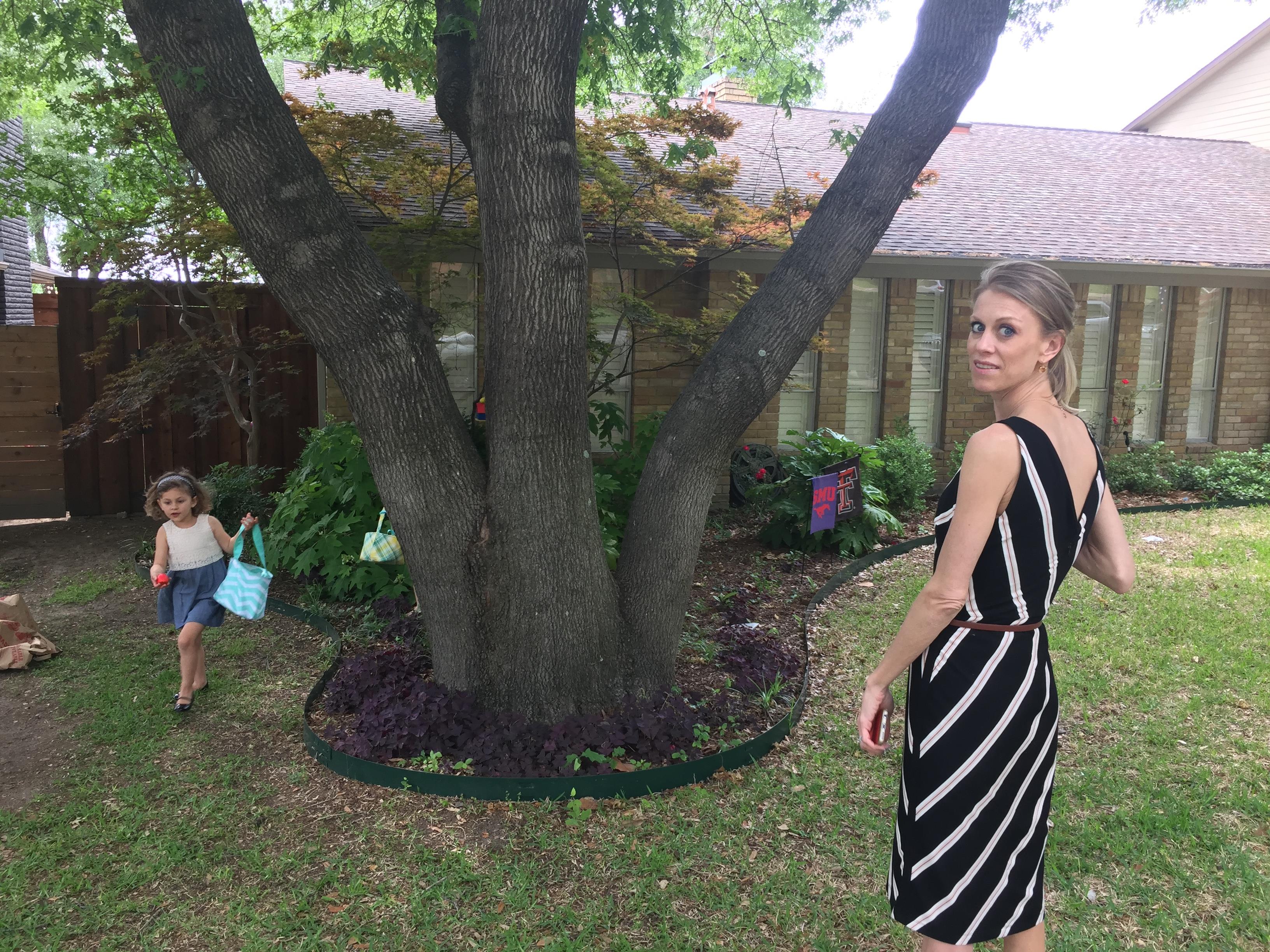 A woman stands near a tree, watching a child play with a bag in a green yard.