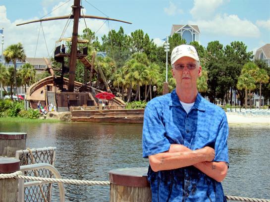 A man poses confidently by the water with a pirate ship attraction in the background.