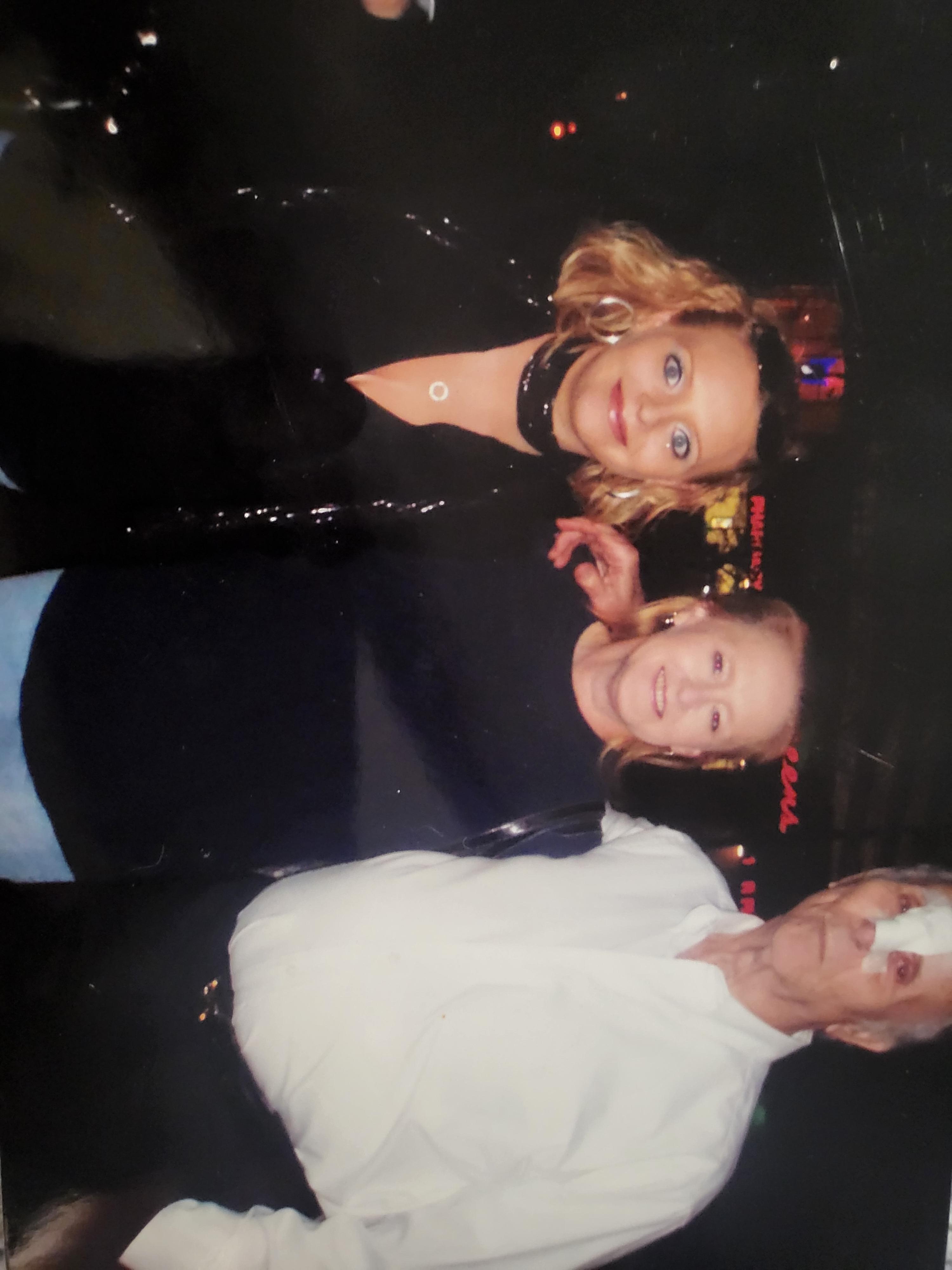 Two women pose happily with a man at a lively evening gathering in a vibrant city.