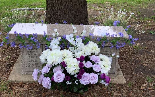 Beautiful floral arrangements decorate a memorial stone under a tree, surrounded by greenery.
