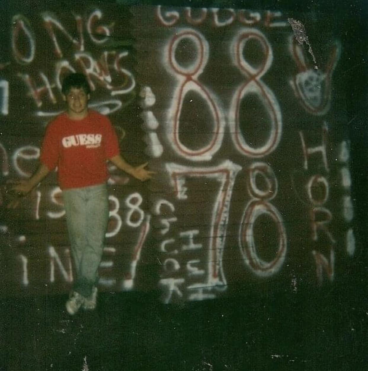 A young person stands playfully with arms outstretched in front of a graffiti wall lit by dim light.