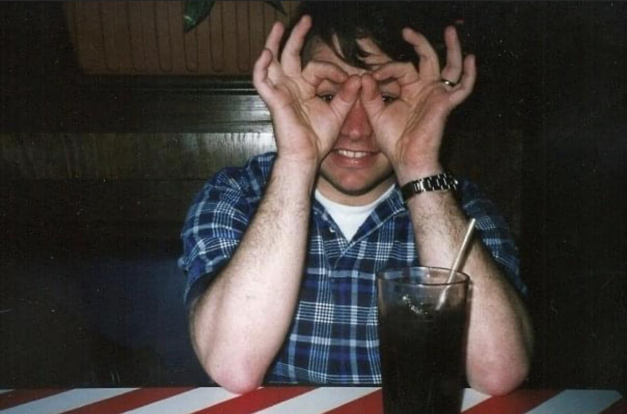 A man playfully creates a silly face by using his hands while enjoying a beverage at a table.