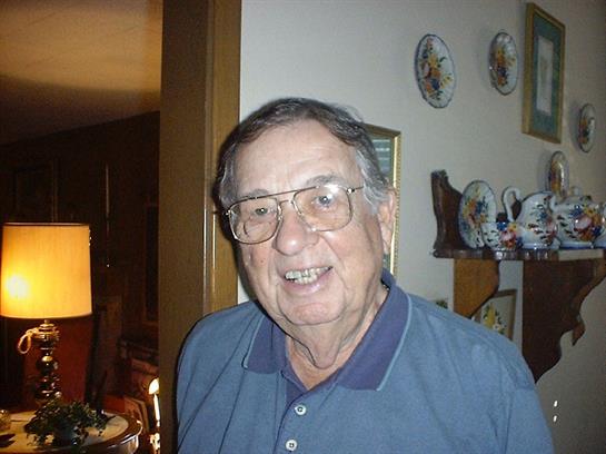 An elderly man wearing glasses smiles warmly in a well-decorated indoor space.