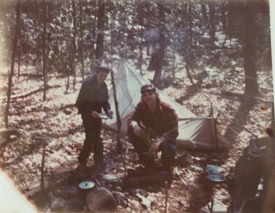 Two individuals are cooking over a fire in a forested area, surrounded by trees and a tent.